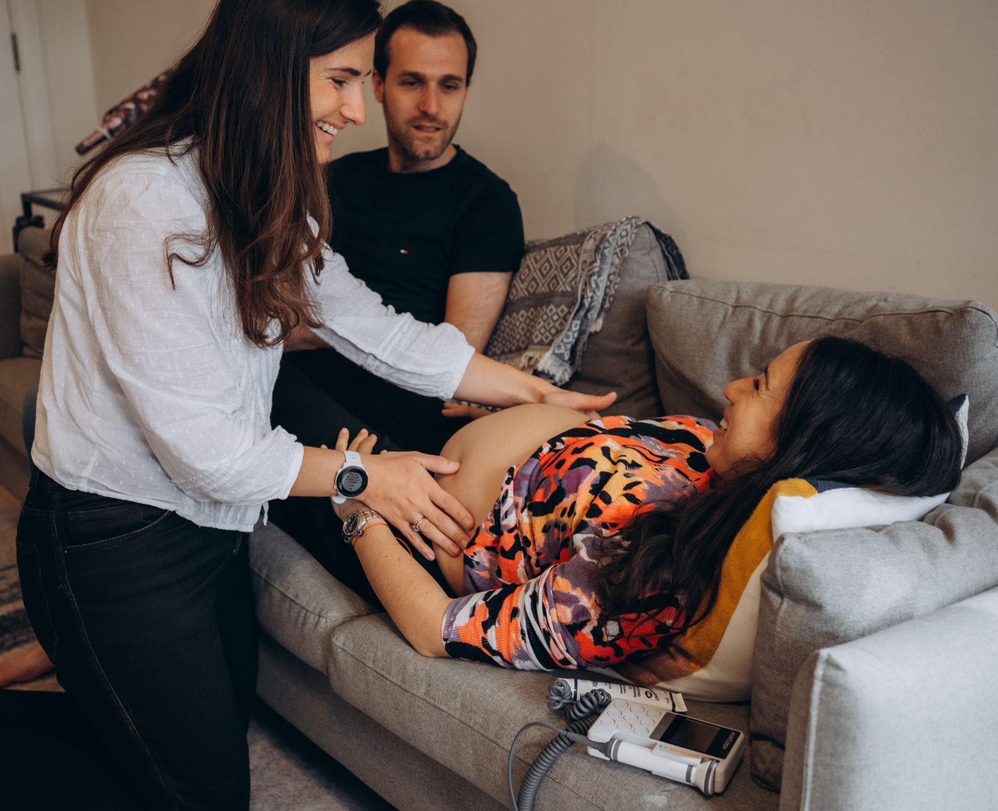 Clarissa, our co-founder and midwife, smiles as she feels a pregnant woman's bump at home. The woman lies laughing on the sofa, her partner beside her.