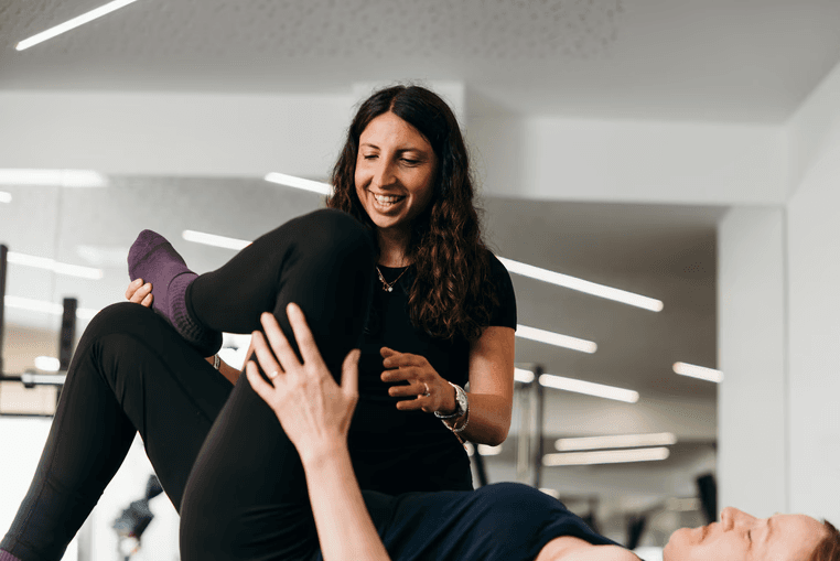 A smiling physio guiding a woman's leg through a stretch as she lies on a mat